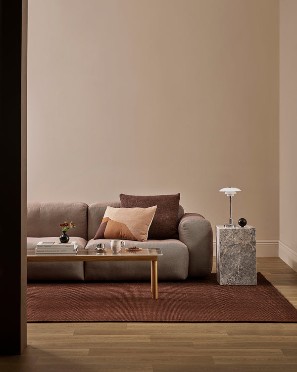 The Baya Linden Floor Rug in Brick styled in a living room beneath a sofa and coffee table, showing its warm red-brown tone against the timber floor.