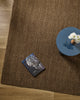 Overhead styling shot of the Baya Linden Floor Rug in Ironbark beside a round side table with a book and a vase of white chrysanthemums.
