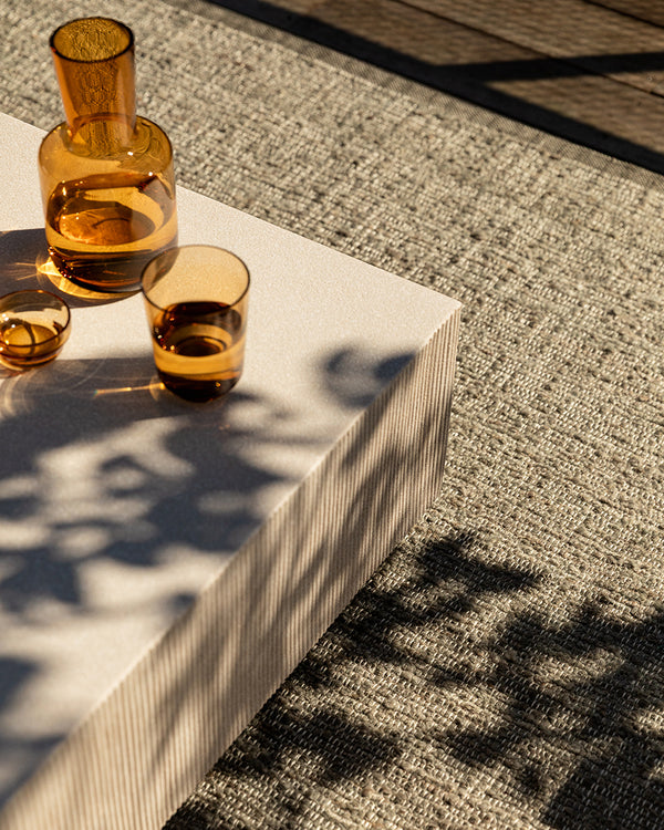 Baya Ormond outdoor rug in Mineral, styled on a deck under a pale stone table with an amber glass carafe and tumblers.
