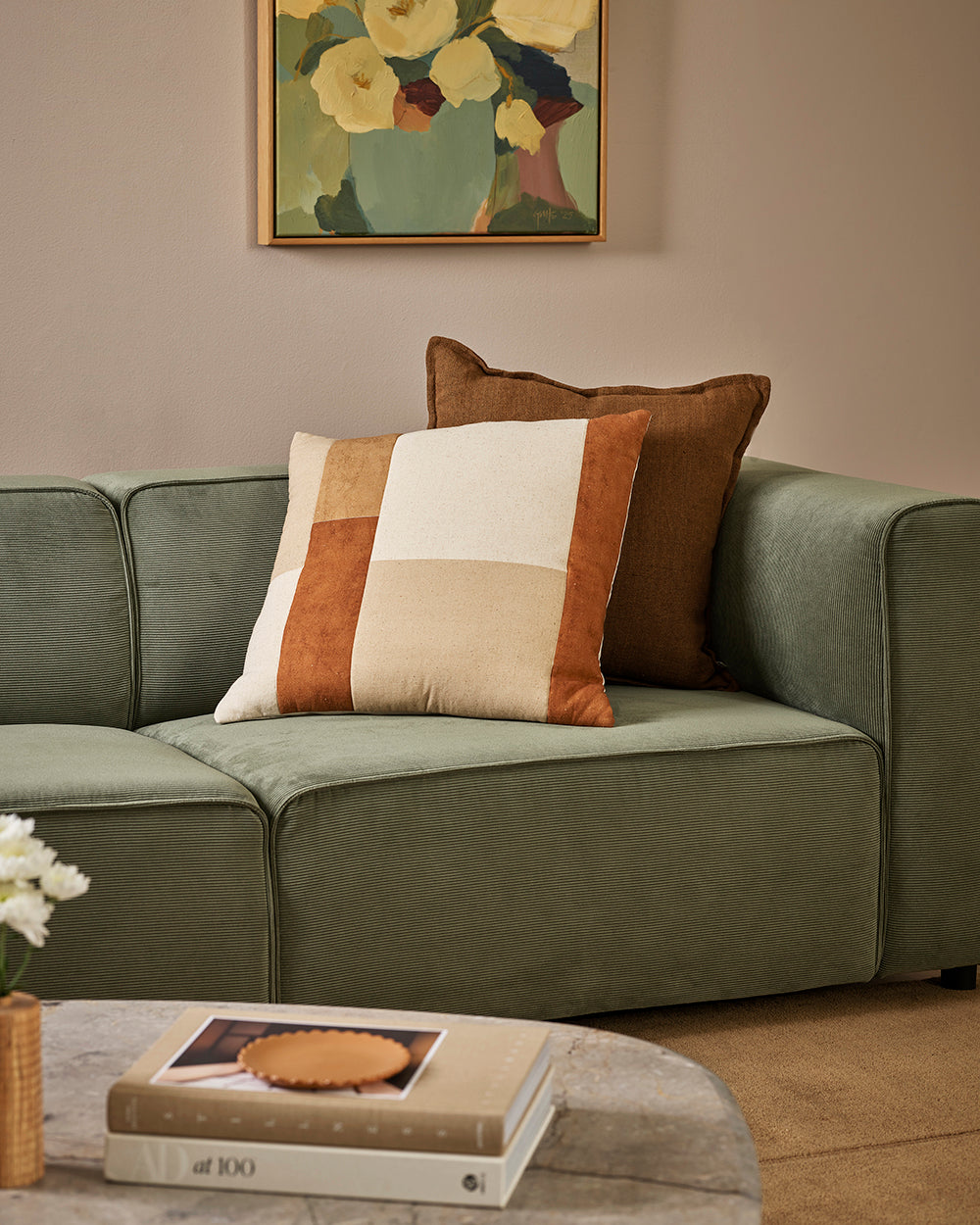 Baya Piazza Cushion in Multi, styled on a sage green sofa in front of a brown cushion, with books on a table.