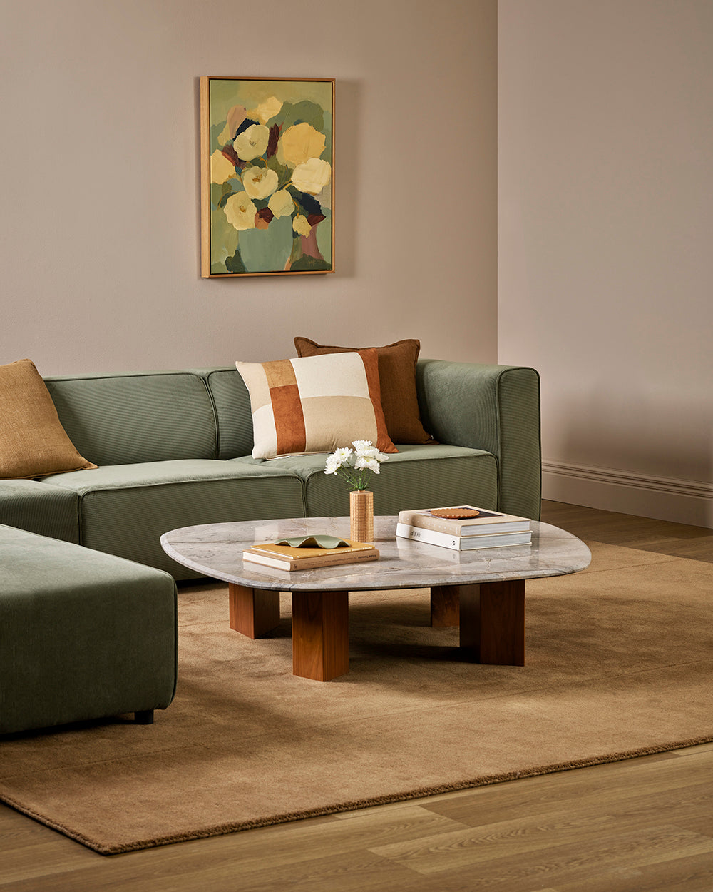 Baya Piazza Cushion in Multi, styled on a sage green sofa in a room with a floral artwork and marble coffee table.