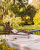 Baya Solar Outdoor Lumbar Cushion in Chocolate, styled on a woven chaise lounge beside a pool.