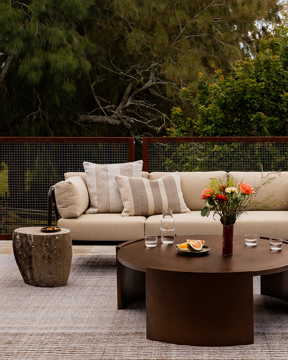 Baya Solar Outdoor Lumbar Cushion in Chocolate, styled on an outdoor sofa beside a round table with flowers and glasses.