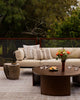 Baya Solar Outdoor Lumbar Cushion in Chocolate, styled on an outdoor sofa beside a round table with flowers and glasses.