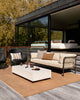 Baya Solar Outdoor Lumbar Cushion in Chocolate, styled on an outdoor sofa in a garden setting with a wicker chair nearby.