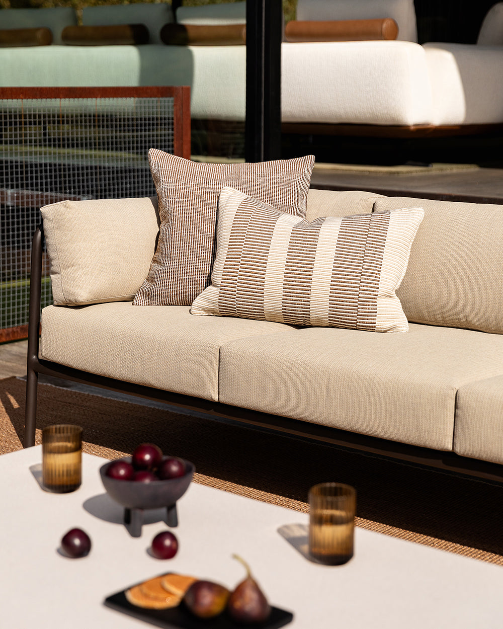 Baya Solar Outdoor Lumbar Cushion in Chocolate, styled on an outdoor sofa beside a plain cushion, with a table setting nearby.