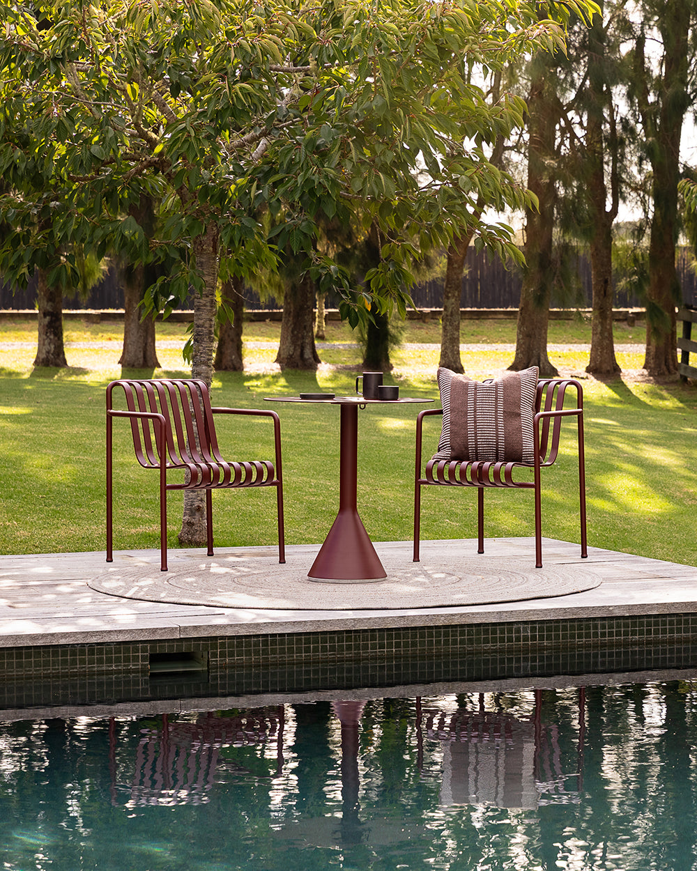 Baya Swell Outdoor Cushion in Chocolate, styled on an outdoor chair beside a pool, with a bistro table and lawn behind.