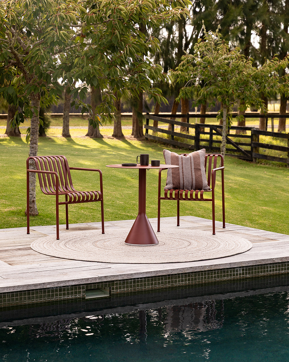 Baya Swell Outdoor Cushion in Chocolate, styled poolside on an outdoor chair beside a bistro table.