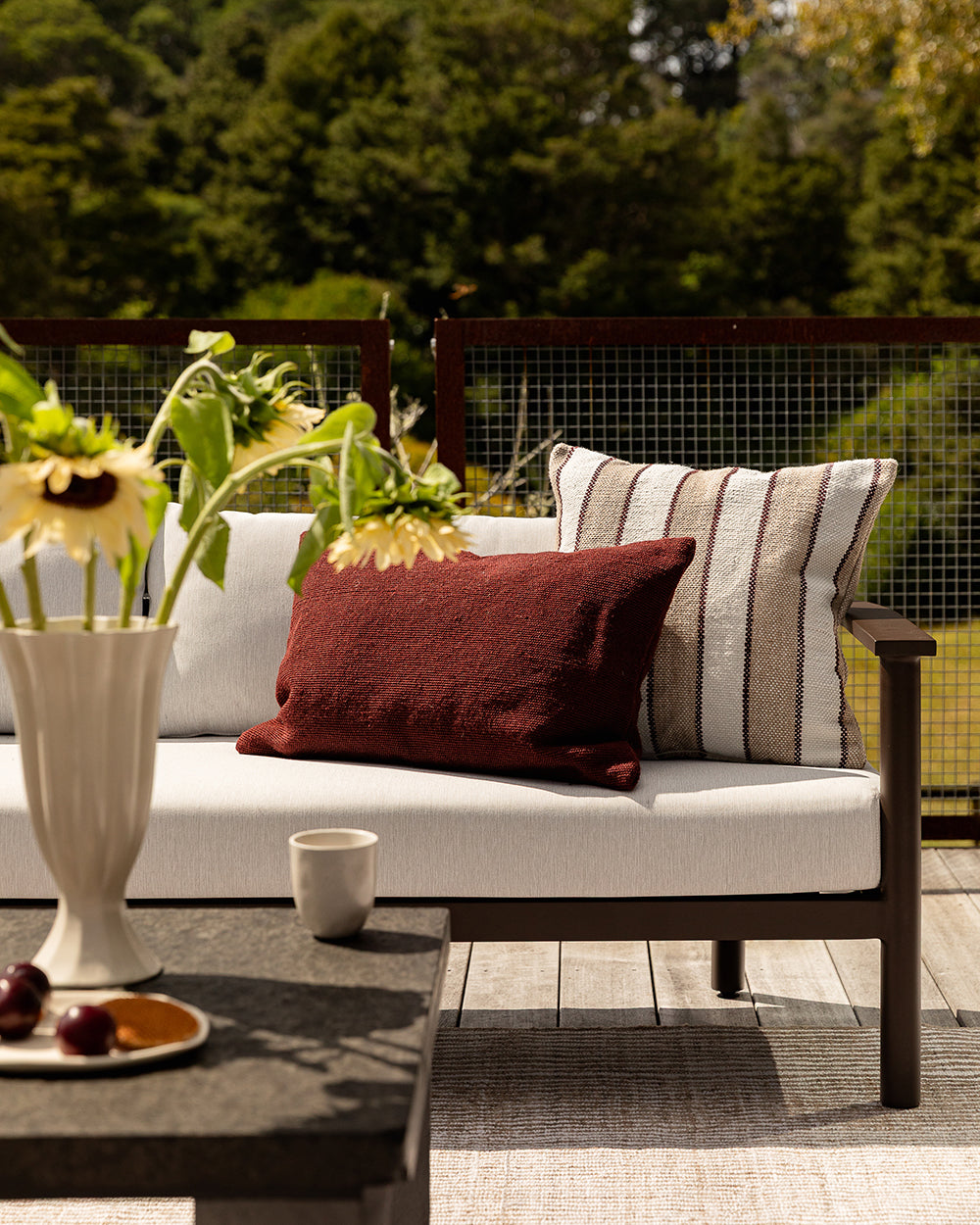 Baya Tidal Outdoor Cushion in Terracotta, styled on an outdoor sofa beside another cushion, with a table setting nearby.