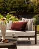 Baya Tidal Outdoor Cushion in Terracotta, styled on an outdoor sofa beside another cushion, with a table setting nearby.