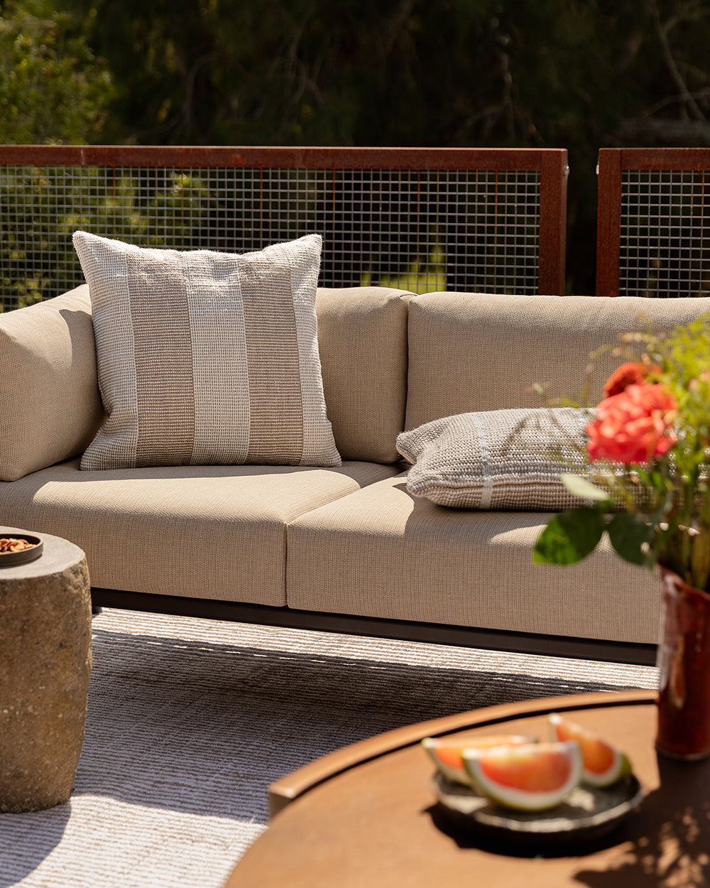 Baya Tide Outdoor Cushion in Almond styled on an outdoor sofa with a grey patterned cushion, flowers and fruit.