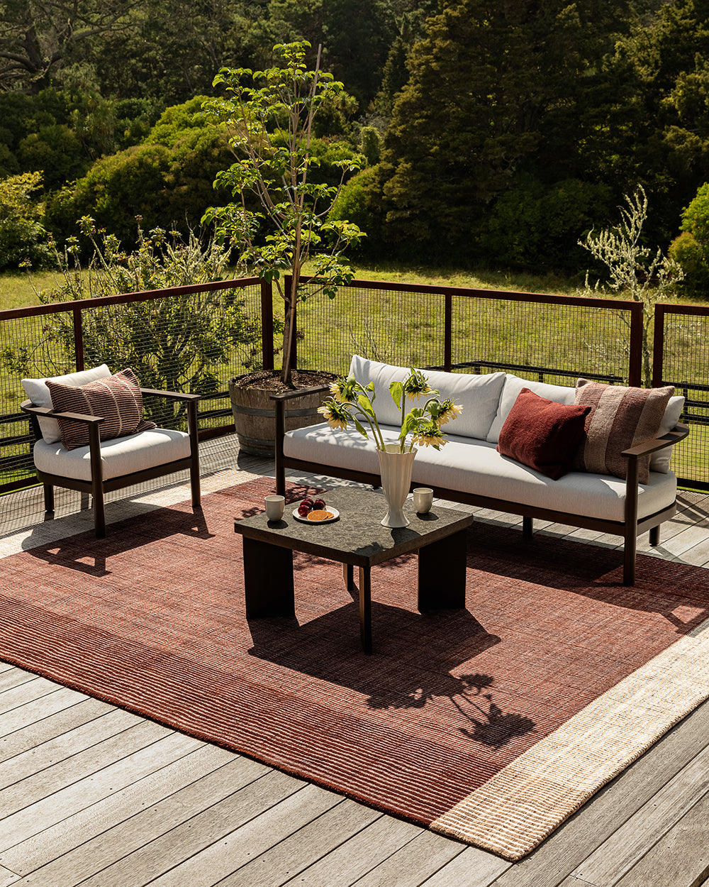Baya Tide Outdoor Cushion in Claret styled on an outdoor sofa and armchair, with sunflowers and a burgundy cushion.
