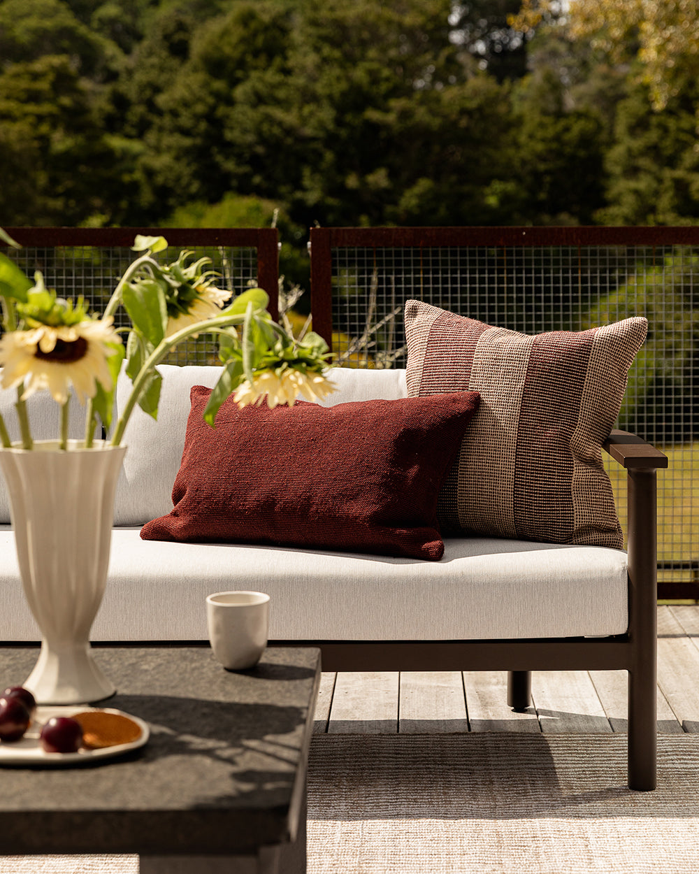 Baya Tide Outdoor Cushion in Claret styled on an outdoor sofa, a closer view with sunflowers and a burgundy cushion.