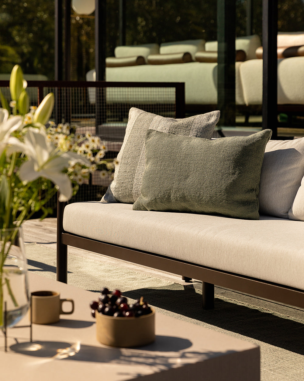 Baya Tide Outdoor Cushion in Eucalyptus styled on an outdoor sofa, a closer view with white lilies in the foreground.