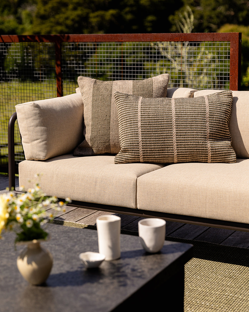Baya Tide Outdoor Cushion in Mangrove styled on an outdoor sofa with a solid dark cushion, flowers and cups.