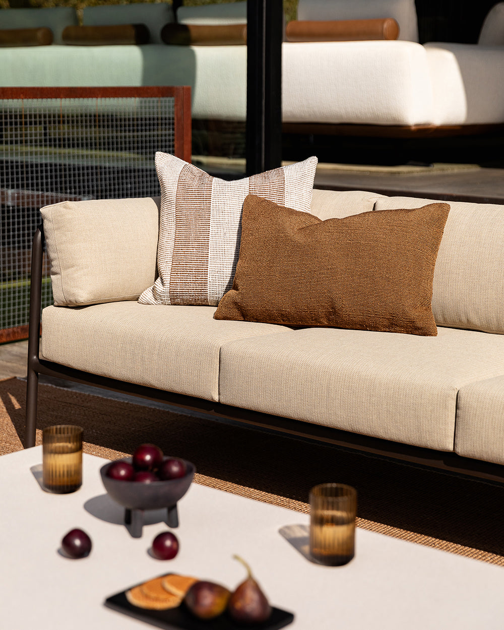 Baya Tide Outdoor Cushion in Tobacco styled on an outdoor sofa with a solid brown Baya cushion.