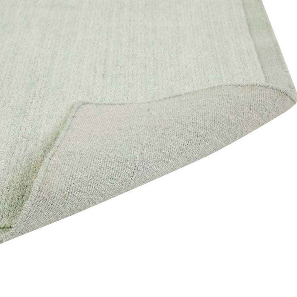 Close up of the corner of a handmade, 100% wool floor rug by Soren Liv NZ and Globe West, in subtle shades of light blue and green, shades of seafoam. The edges of the rug are indented, lending subtle sophistication, and the corner is folded over to view the underside of the rug. 