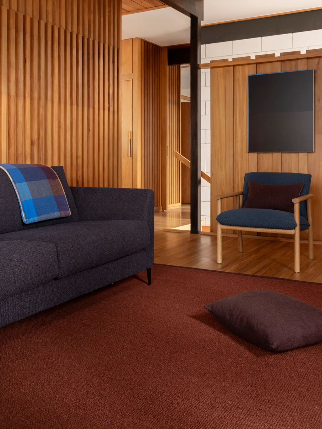 Città Joni Wool Mat in Paprika/Mulberry, styled in a wood-panelled living space with a sofa and armchair.