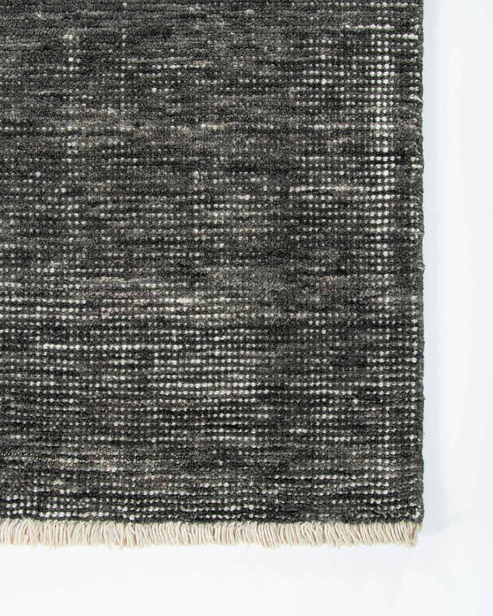 A corner image from above of the Corbett floor rug in the shade Peat, a lightly distressed contemporary charcoal, with cream detailing and a short creamy fringe. By Baya