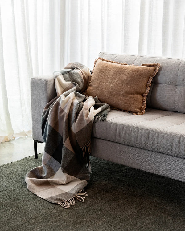 Tan brown lumbar cushion with soft fringe detail decorating a couch alongside a plaid throw blanket