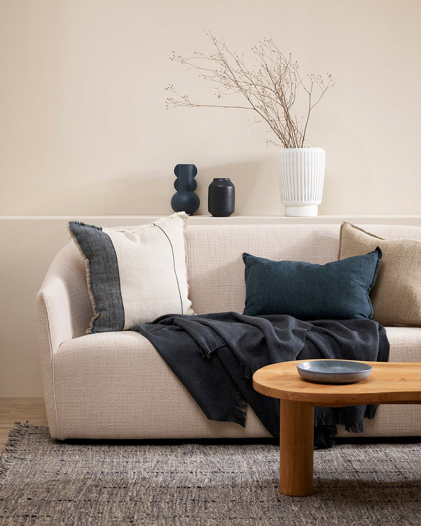 A natural linen cushion with designer stripe and fringed edge detail, shown on a couch with blue cushion and throw