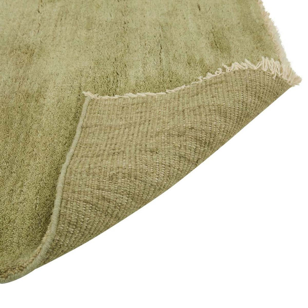 Corner image of a soft leaf-green handloomed woolen rug by Soren Liv. The edges are fringed with wool in a creamy ivory and flipped up to reveal the underside.