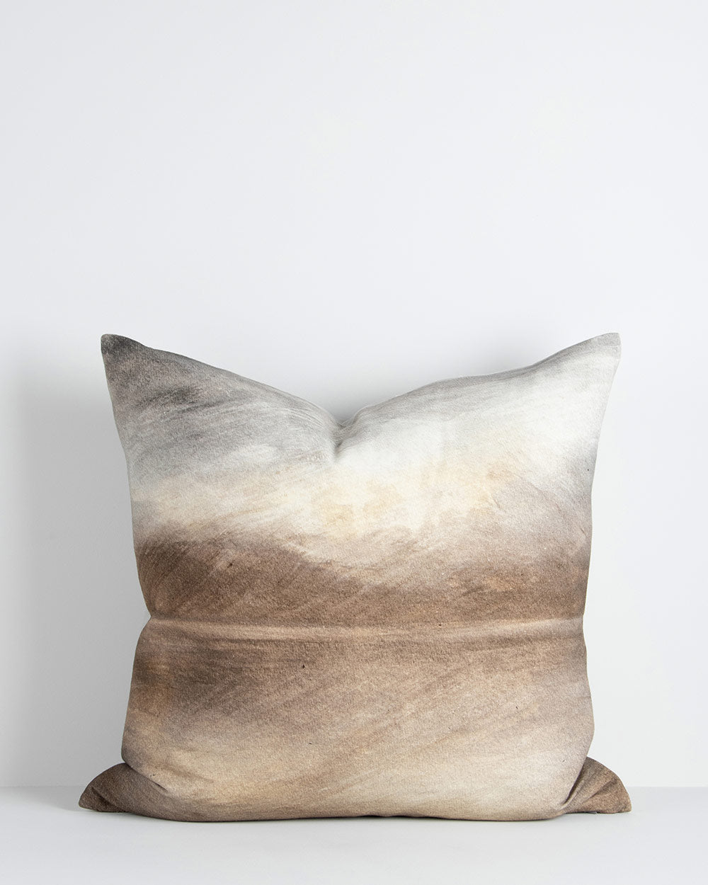 A printed cushion depicting an abstract watercolour landscape scene in shades of sienna, umber and grey. By Baya