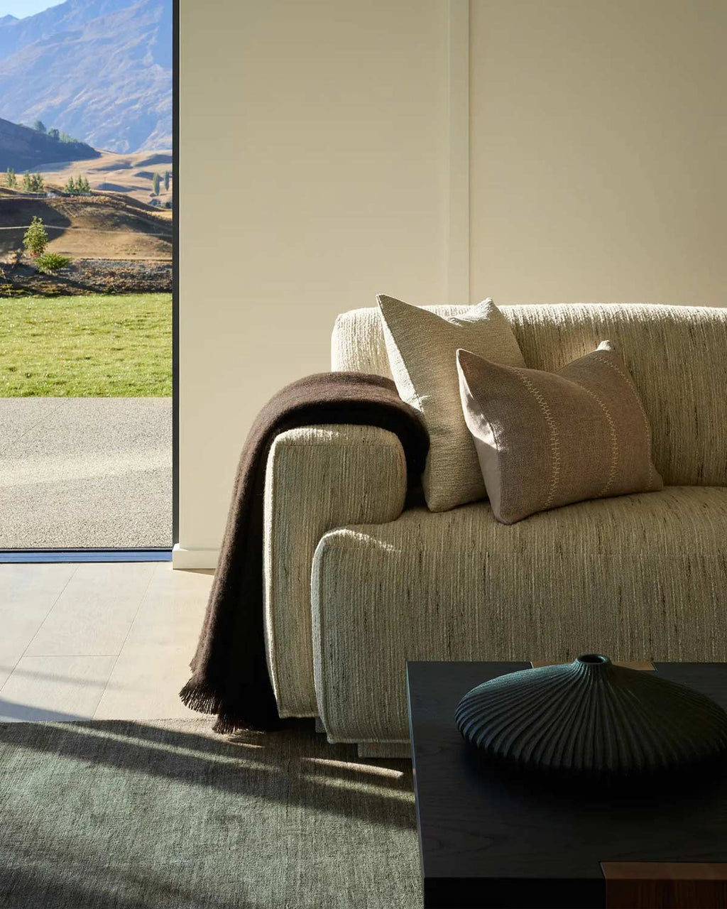 A deep chocolate brown throw blanket draped over the side of a couch. The sun hits it, showing the warm tones in the wool fibres.