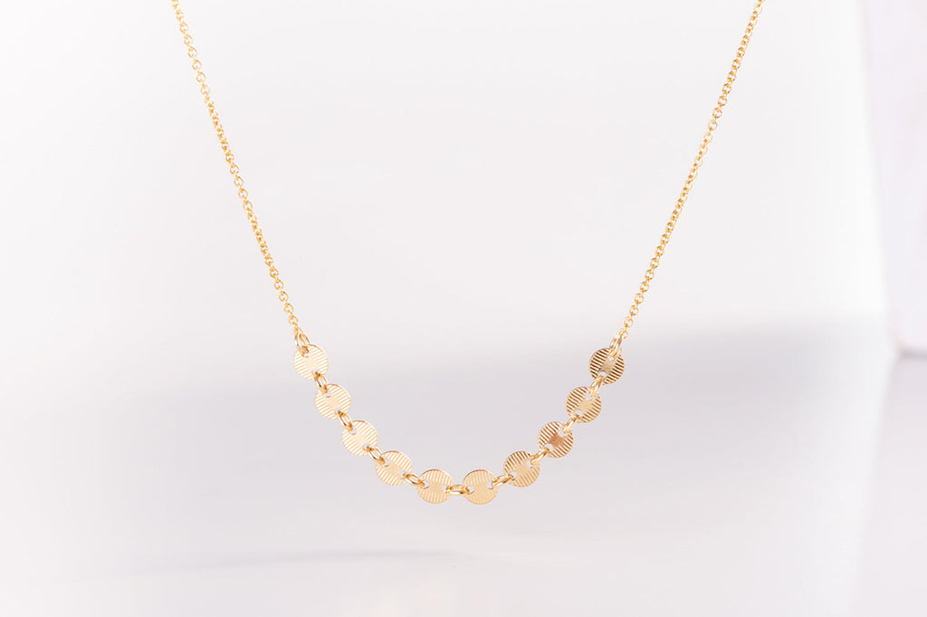 A close up view of the Italy necklace by Zoaje. Ten brushed metal gold-filled discs lie along the middle of the necklace. 