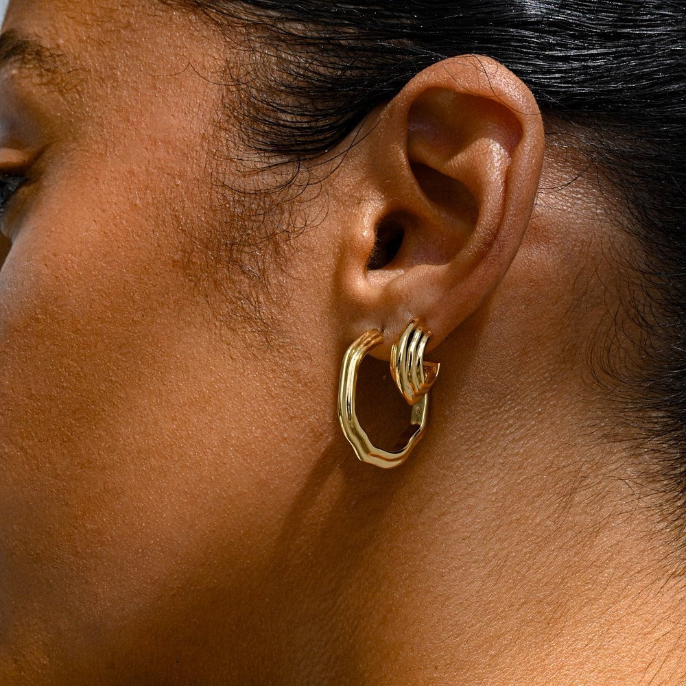 A woman wearing two gold-plated hoop earrings with sculptural detailing. 