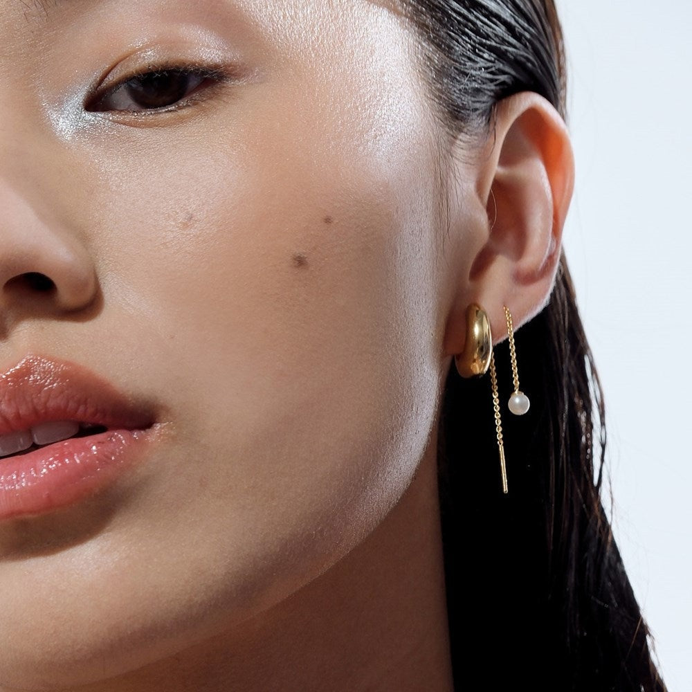 A woman wearing a chunky gold huggie hoop earring, paired with a gold chain drop earring. 