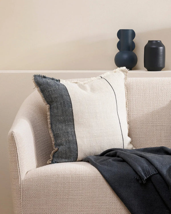 A natural linen cushion with designer stripe and fringed edge detail, shown on a couch with a navy blue throw