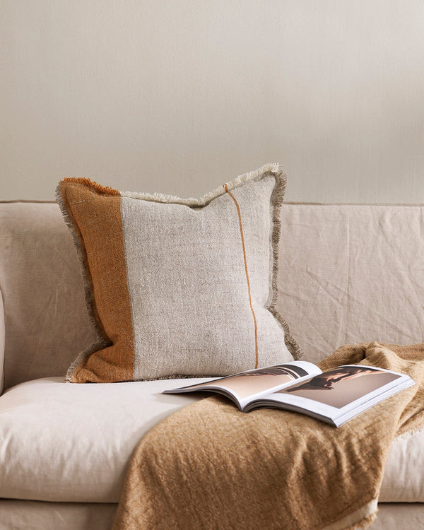 A natural linen cushion with a rust-coloured stripe and fringe, seen on a couch with a throw and magazine