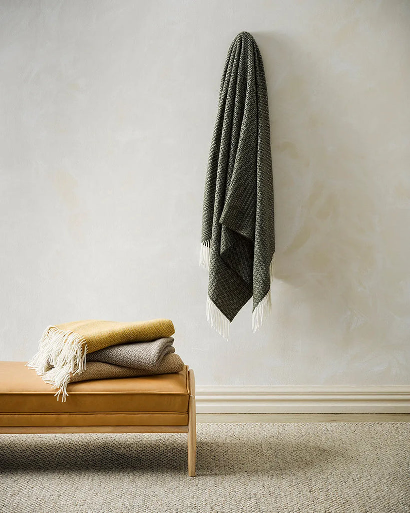 The soft, Baya 'Kale' dark green wool throw blanket hanging from a hook on the wall