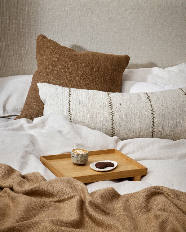 An extra-long lumbar, textural cotton cushion featuring a delicate Cretan-stitch is embroidered in khaki over a natural, ecru-toned base, on a cream and brown bed