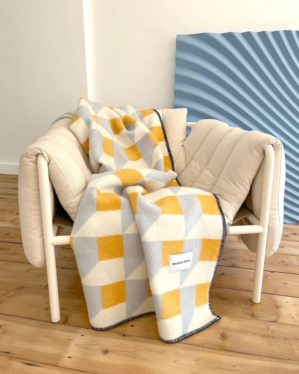 Maison Deux Cubics wool throw in Blue Honey, draped over a cream armchair against a blue ribbed wall panel