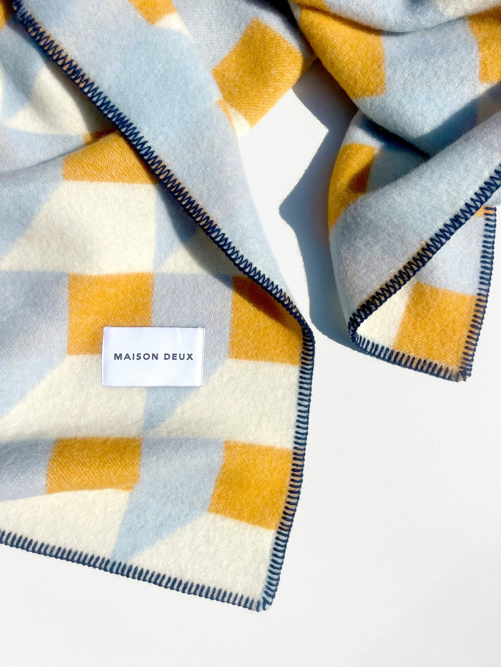 Maison Deux Cubics wool throw in Blue Honey, folded corner detail showing the navy blanket stitched edge and the woven Maison Deux label