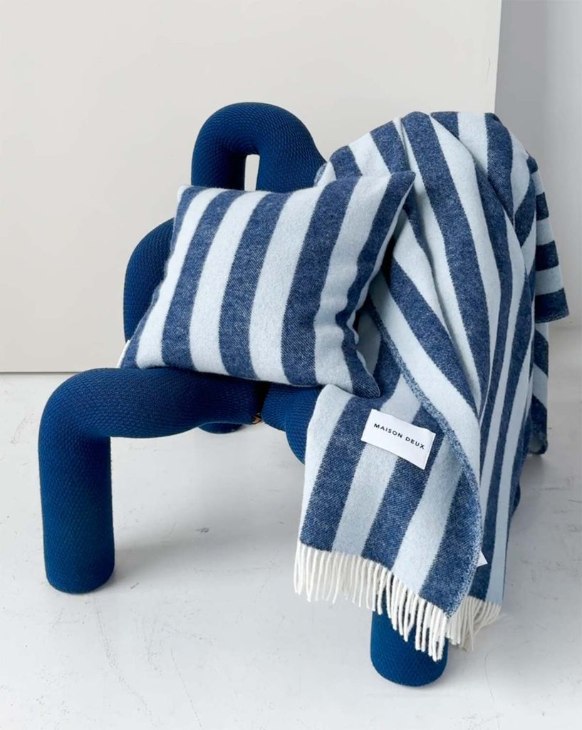 A modern designer blue chair with a matching cushion and throw blanket on it, both striped in shades of navy and light blue. 
