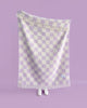 A figure holding up an oversized throw blanket with a stylish checkerboard pattern in shades of pale purple and creamy white