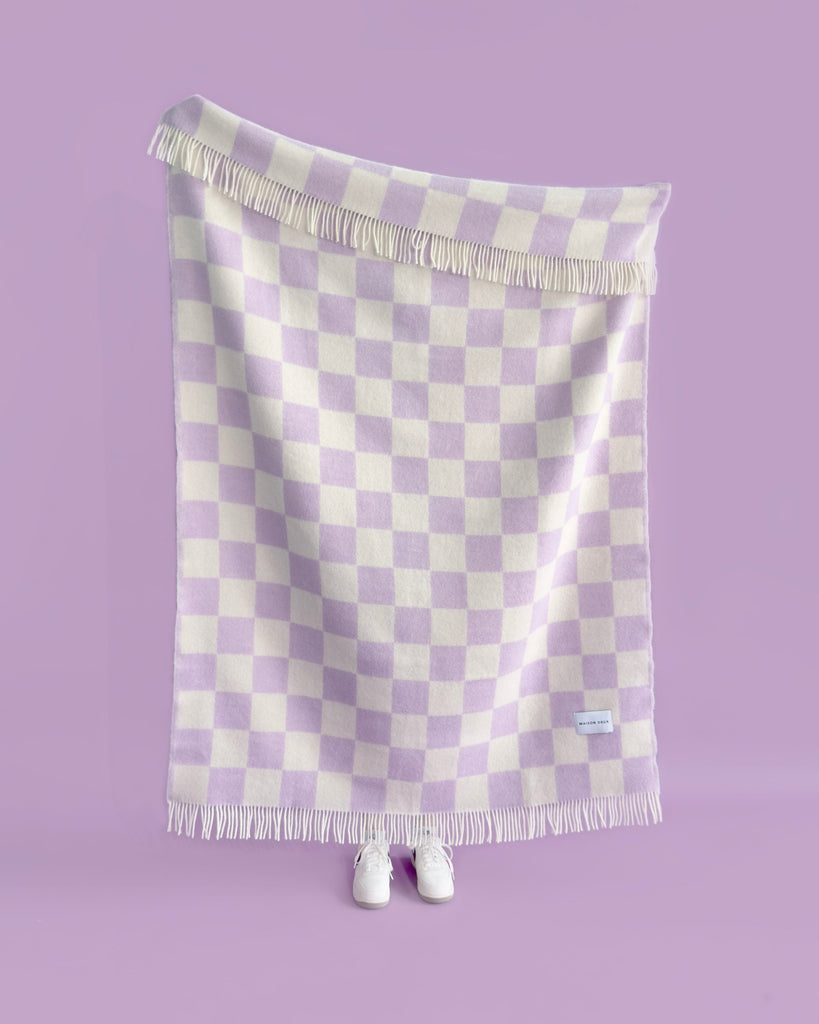 A figure holding up an oversized throw blanket with a stylish checkerboard pattern in shades of pale purple and creamy white