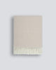 A neatly folded beige 100% merino wool throw blanket with a soft texture and a delicate cream fringe along the edge, by Baya