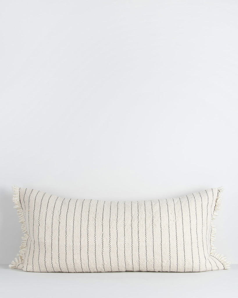 A lengthy cotton lumbar cushion, with regular vertical blue stripes across a cream background. The cushion is fringed at the edges, which adds softness and sophistication. By Baya
