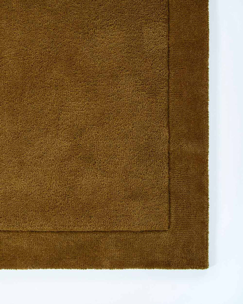 The corner of the Norfolk floor rug in the shade Brass. The border of the rug is cut-pile in a minimalist and refined design. 
