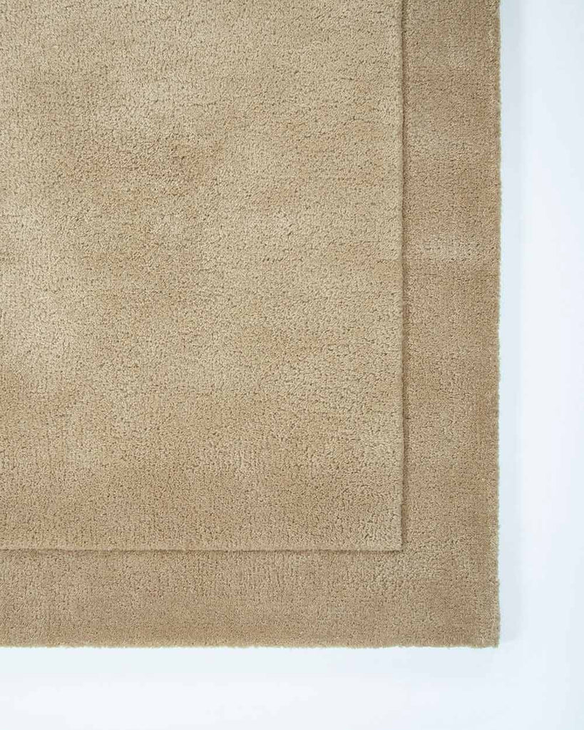 The corner of the Norfolk floor rug in the shade Sand. The border of the rug is cut-pile in a minimalist and refined design. 
