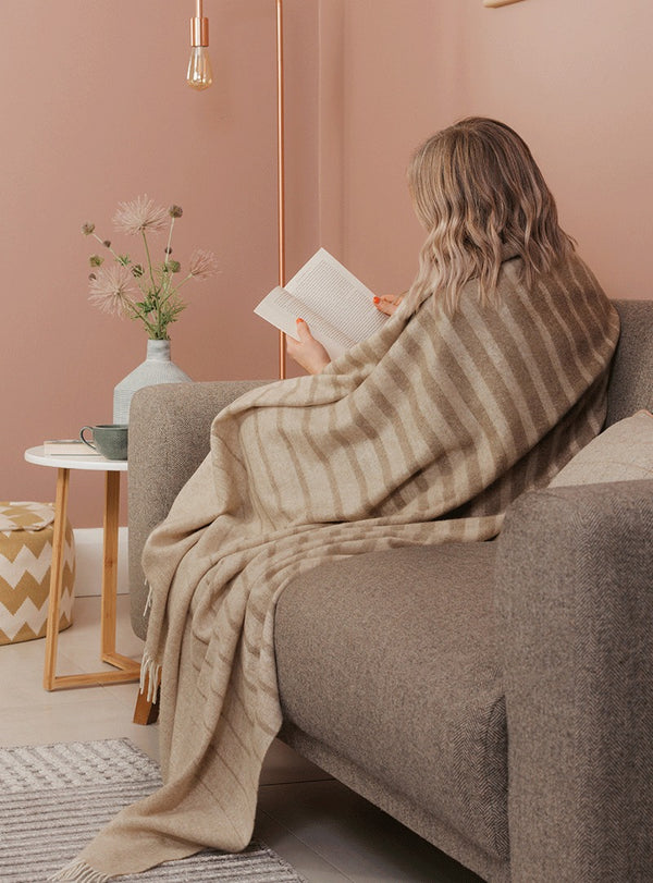NZ merino wool throw blanket featuring a cream and beige stripe design, seen wrapped around a lady in her home