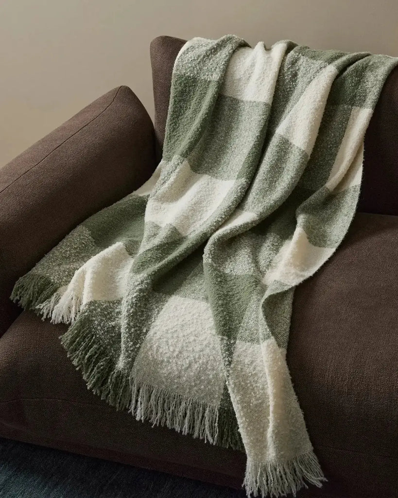 A green and white boucle wool throw blanket with fringe draped over a brown couch