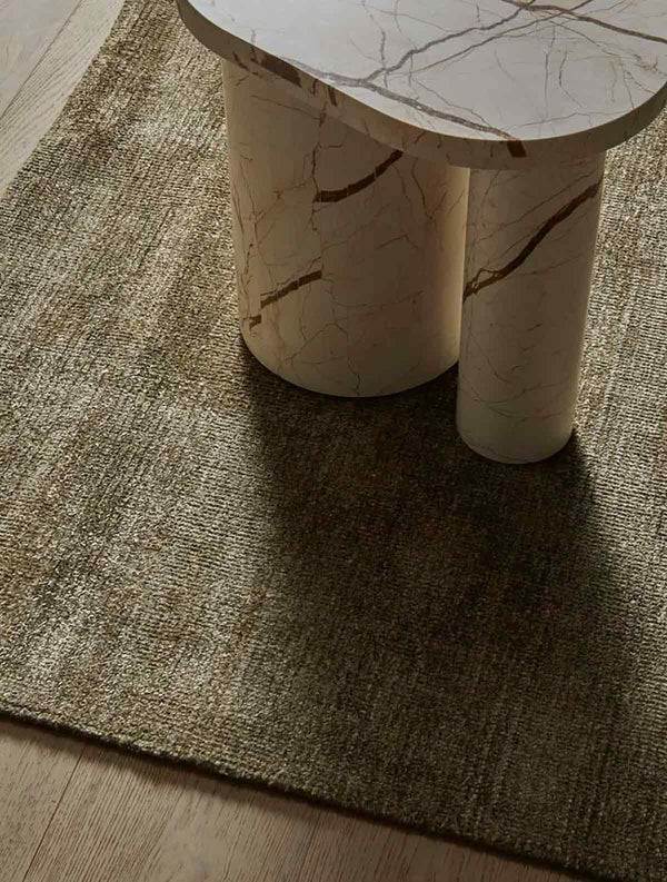 A soft, wool blend rug in shades of muted olive green by Soren Liv NZ and Globe West, on a wooden floor underneath a designer coffee table