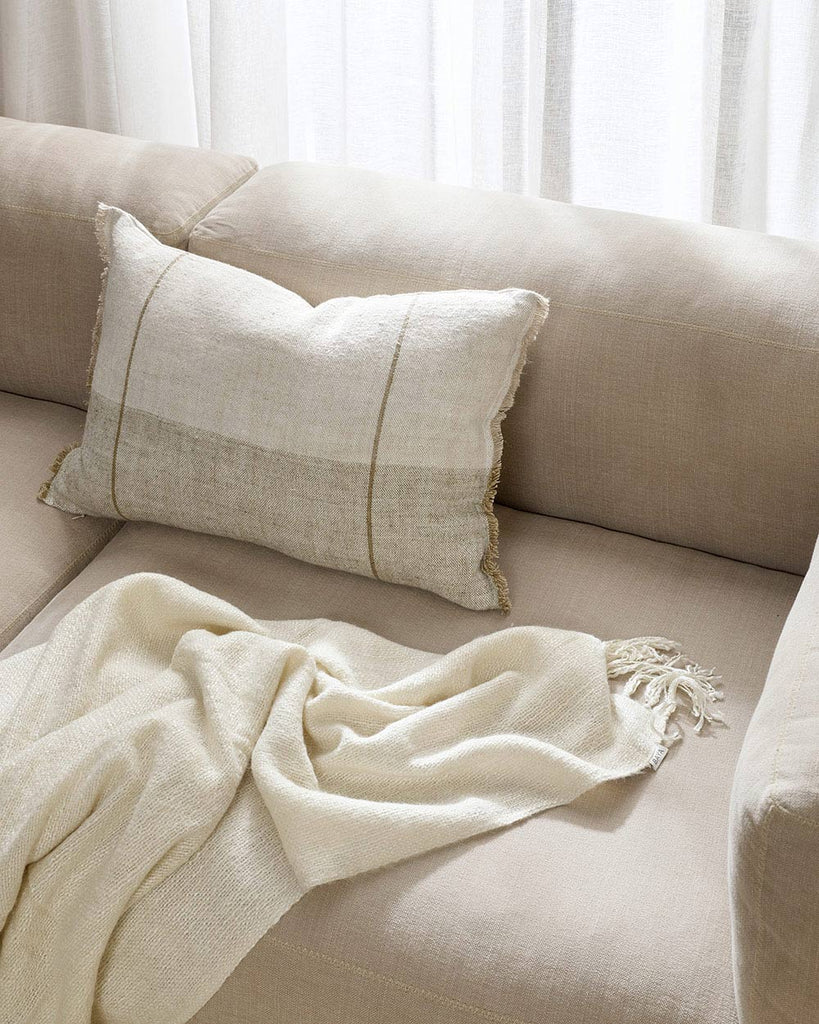 A stylish lumbar cushion in tones of sand beige and cream, with a soft off-white throw blanket on a beige couch by Baya. 