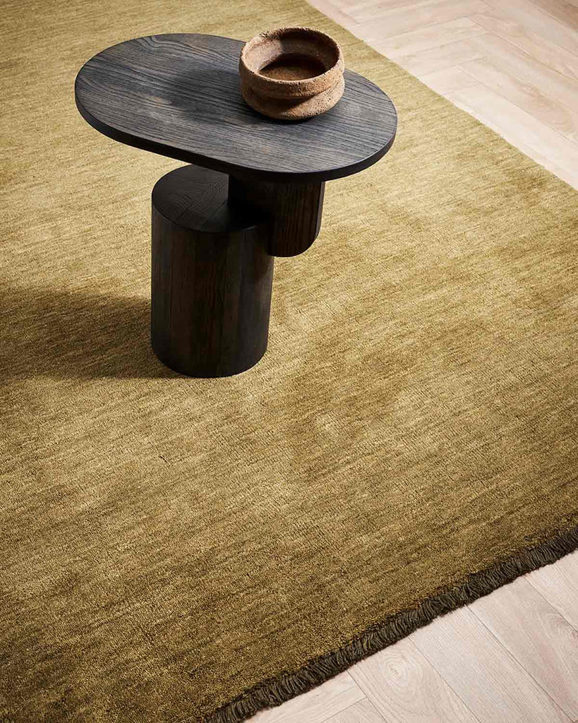 A designer coffee table atop a warm olive-green floor rug in 100% wool with fringed edges. 
