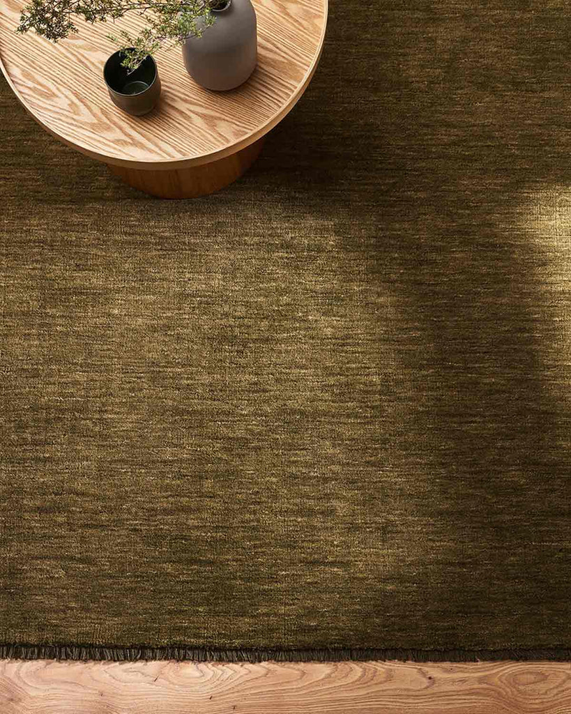 A floor rug in an on trend, warm green tone with fringed edges seen from above with a chic light-wood coffee table. 